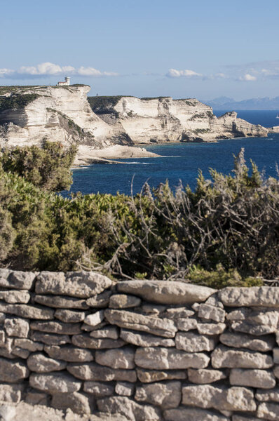 Корсика: каменная стена и захватывающие дух белые известняковые скалы Бонифачо в Международном морском парке Буш-де-Бонифачо, природном заповеднике, созданном в 1993 году, с видом на маяк мыса Пертусато
