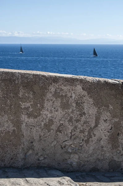 Corsica: stone wall and sailboats sailing in the Strait of Bonifacio, the stretch of sea between Corsica and Sardinia which divides the Tyrrhenian Sea from the western Mediterranean Sea