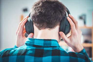 Mavi gömlekli genç beyaz adam kablosuz kulaklıkla müzik dinliyor. Başının arkasına..