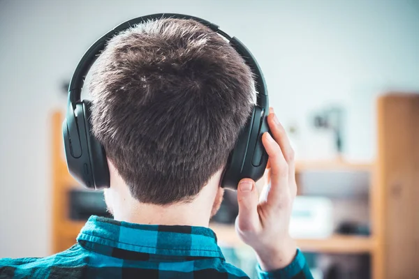 Young Caucasian man with checked blue shirt is listing to music with wireless headphones. Back of the head.