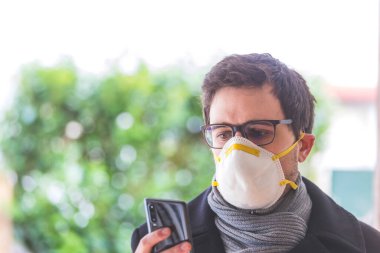 Young business man outdoors with a breathing mask and smartphone. Flue and corona season.  clipart