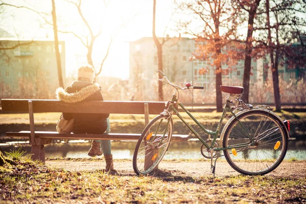 公園のベンチで太陽を楽しんでいる自転車と若い女性のバックビュー 春の時間 — ストック写真