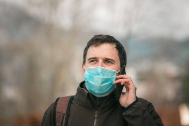 Açık havada maske takan ve akıllı telefon kullanan genç bir adam. Corona virüsü ve grip mevsimi.