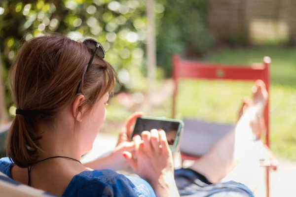 Girl is sitting on the sunny porch, enjoying the day and using her smartphone
