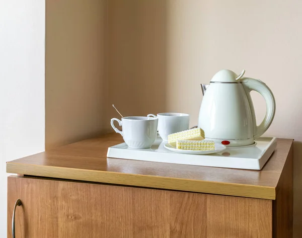 Tray with electric kettle and cups are on wooden low cabinet. Fragment of interior. Copy space