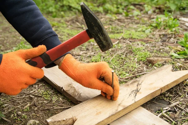 Hands in orange gloves hammer a nail with a red handle into a board with a red handle