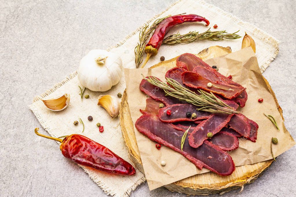 Basturma, dried tenderloin of beef meat, jerky, thinly sliced. Dry rosemary, pepper mix, chili, garlic on vintage linen cloth. Delicious food on a wooden board, stone background, close up.