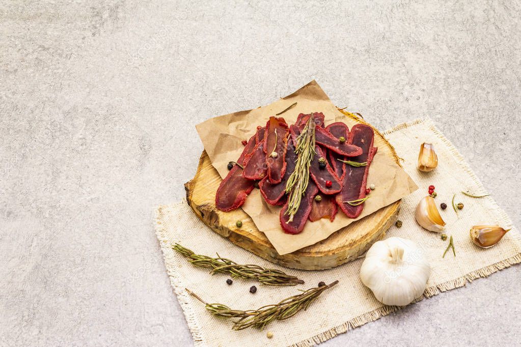 Basturma, dried tenderloin of beef meat, jerky, thinly sliced. Dry rosemary, pepper mix, chili, garlic on vintage linen cloth. Delicious food on a wooden board, stone background, copy space.