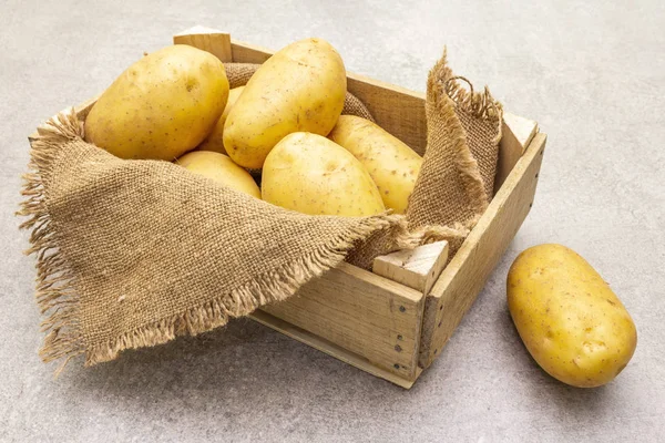Batatas jovens cruas não descascadas em uma caixa de madeira. Nova colheita, em pano de saco, fundo de pedra, close . — Fotografia de Stock