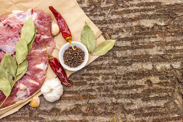 Raw pork shoulder with spices. Bay leaf, garlic, chili. On a wooden bark background, close up, top view, copy space.