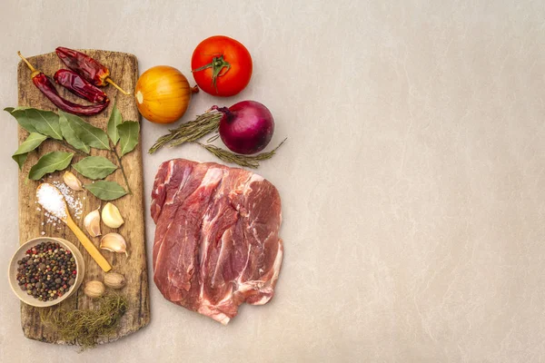 Raw pork shoulder. Spices, dry herbs, onions, garlic, rosemary, bay leaf, hot pepper, nutmeg, pepper mix, tomatoes. Ingredients for marinating meat. Wooden vintage board, stone background, top view.