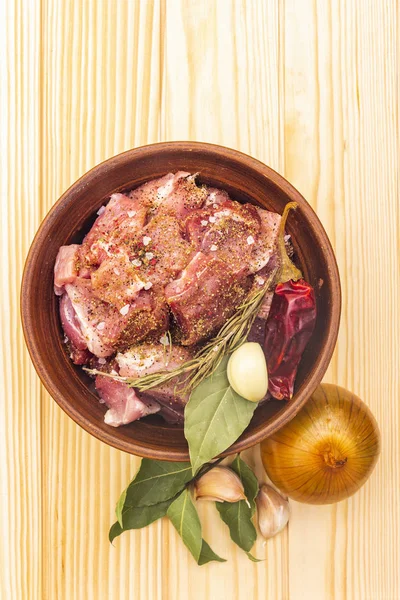 Raw pork shoulder. Spices, dry herbs, garlic, rosemary, bay leaf, hot pepper, nutmeg, pepper mix, onion. Ingredients for marinating meat. Rustic ceramic bowl on wooden background, top view, close up.