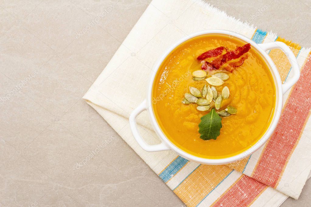 Pumpkin cream soup. White ceramic bowl with fresh pumpkin, dry seeds, smoked bacon. With vintage linen cloth (napkin) on stone background, top view