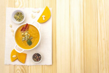 Balkabağı kremalı çorba. Taze kabaklı beyaz seramik kase, kuru kabak çekirdekleri, tütsülenmiş pastırma, bitki. Ahşap zemin üzerinde kaliteli keten kumaş (peçete), üst görünüm