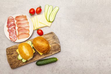 Hamburger ekmeği, adım adım hazırlık. Salam, kurutulmuş domuz eti, domates, salatalık, peynir malzemeleri. Tahta ve taş zemin üzerinde, üst görünüm.