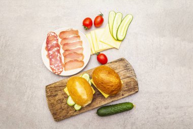 Hamburger ekmeği, adım adım hazırlık. Salam, kurutulmuş domuz eti, domates, salatalık, peynir malzemeleri. Tahta ve taş zemin üzerinde, üst görünüm.