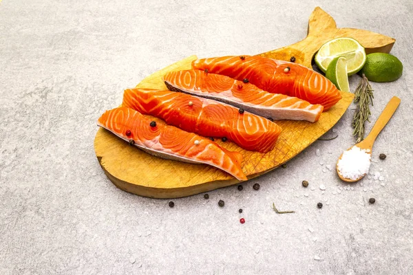 Raw trout (salmon) piece with lime, rosemary, sea salt and pepper mix. Wooden board in the shape of a fish, on a stone background. — Stock Photo, Image