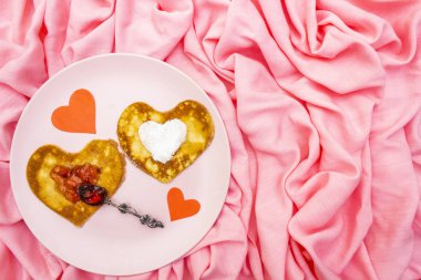 Çilek reçeli, gümüş kaşık ve kağıt kalpli romantik kahvaltı için kalp şeklinde krepler. Shrovetide (karnaval) konsepti. Pembe kumaş arkaplanda, üst görünümde.