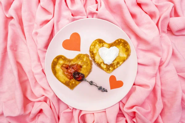 Crêpes en forme de coeur pour le petit déjeuner romantique avec confiture de fraises, cuillère en argent et coeurs en papier. Concept de Shrovetide (carnaval). Sur fond lin tissu rose, vue de dessus — Photo