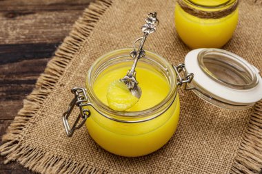 Pure or desi ghee (ghi), clarified melted butter. Healthy fats bulletproof diet concept or paleo style plan. Glass jar, silver spoon on vintage sackcloth. Wooden boards background clipart