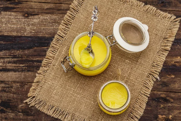 Ghee puro ou desi (ghi), manteiga derretida clarificada. Gorduras saudáveis conceito de dieta à prova de balas ou plano de estilo paleo. Frasco de vidro, colher de prata no saco vintage. Placas de madeira fundo — Fotografia de Stock