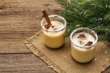 Eski ahşap masada noel süsüsüyle düzenlenmiş iki bardakta yumurtalı kokteyl. Evergreen fer ağaç dalı, yapay kar, çuval bezi