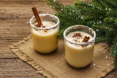 Eski ahşap masada noel süsüsüyle düzenlenmiş iki bardakta yumurtalı kokteyl. Evergreen fer ağaç dalı, yapay kar, çuval bezi