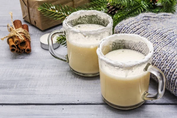 Коктейль Eggnog в двух бокалах кружка устроена с рождественским украшением на светлом деревянном столе. Вечнозеленая елка ветка, подарок, уютная майка, искусственный снег — стоковое фото