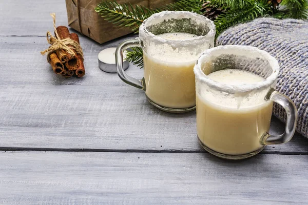 Коктейль Eggnog в двух бокалах кружка устроена с рождественским украшением на светлом деревянном столе. Вечнозеленая елка ветка, подарок, уютная майка, искусственный снег — стоковое фото