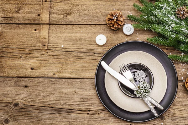 クリスマスと新年のディナーの場所の設定。常緑モミの木の枝、キャンドル、コーン、セラミックプレート、フォークとナイフ。冬のカトラリー木製の背景 — ストック写真