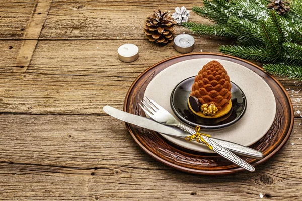 Christmas and New Year dinner place setting. Sweet snack, fir tree branch, candles, cones, ceramic plates, fork and knife. Winter cutlery wooden background