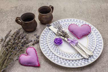 Hafif beton arka planda romantik bir masa. Sevgililer Günü ya da düğün kartı şablonu. Leylak kalp, lavanta, gül çiçeği, çatal bıçak, bardak.