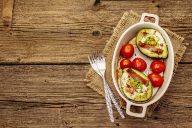 Güneşli sabah kahvaltısı. Avokado yarısı pişmiş yumurta, pastırma, domates, baharatlar. Sağlıklı yiyecekler, ketojenik diyet konsepti. Eski tahta arka plan