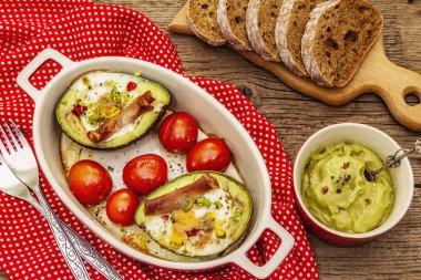 Güneşli sabah kahvaltısı. Avokado yarısı pişmiş yumurta, pastırma, domates, baharatlar. Sağlıklı yiyecekler, ketojenik diyet konsepti. Eski tahta arka plan