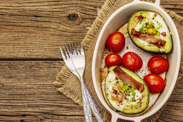 Güneşli sabah kahvaltısı. Avokado yarısı pişmiş yumurta, pastırma, domates, baharatlar. Sağlıklı yiyecekler, ketojenik diyet konsepti. Eski tahta arka plan