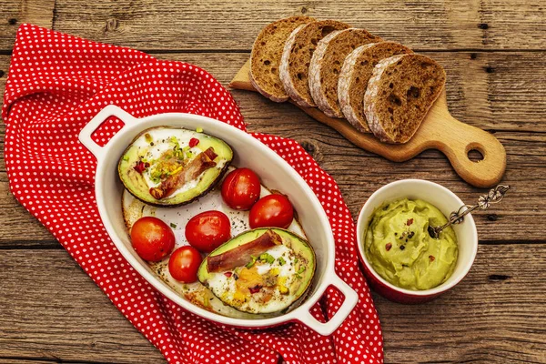 Güneşli sabah kahvaltısı. Avokado yarısı pişmiş yumurta, pastırma, domates, baharatlar. Sağlıklı yiyecekler, ketojenik diyet konsepti. Eski tahta arka plan