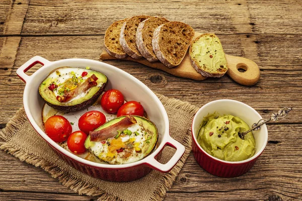 Güneşli sabah kahvaltısı. Avokado yarısı pişmiş yumurta, pastırma, domates, baharatlar. Sağlıklı yiyecekler, ketojenik diyet konsepti. Eski tahta arka plan