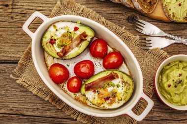 Güneşli sabah kahvaltısı. Avokado yarısı pişmiş yumurta, pastırma, domates, baharatlar. Sağlıklı yiyecekler, ketojenik diyet konsepti. Eski tahta arka plan