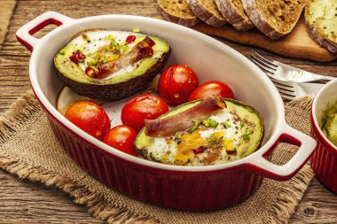 Güneşli sabah kahvaltısı. Avokado yarısı pişmiş yumurta, pastırma, domates, baharatlar. Sağlıklı yiyecekler, ketojenik diyet konsepti. Eski tahta arka plan