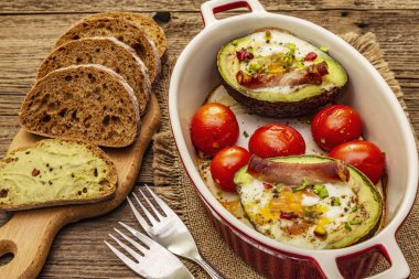 Güneşli sabah kahvaltısı. Avokado yarısı pişmiş yumurta, pastırma, domates, baharatlar. Sağlıklı yiyecekler, ketojenik diyet konsepti. Eski tahta arka plan