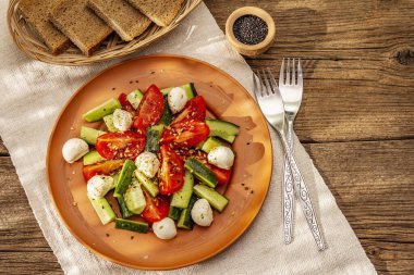 Domates, salatalık ve bebek mozzarellasından taze salata. Kabuklu ekmekle güneşli bir öğle yemeği. Eski tahta arka plan, üst görünüm