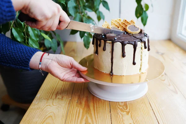 Woman\'s hands cut the cake with caramel cream and chocolate ganache on wooden table.