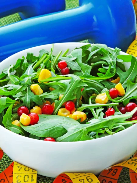 Bright fitness salad with arugula corn and cowberry in a white bowl. Dumbbells and measuring tape. The concept of weight loss and nutrition. Tasty and healthy diet salad.