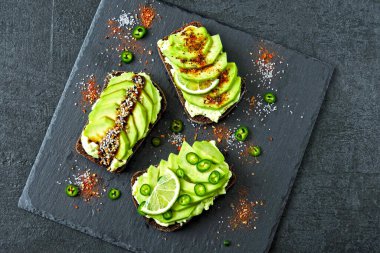 Lezzetli malzemelerle kaplanmış avokado tostu. Avokado tostları servis için gri bir tahtada sunuluyor..