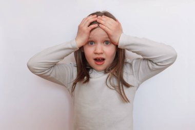 Açık renk monokrom kazaklı, elleri beyaz gri bir arka plana bağlanmış ve kameraya bakan küçük üzgün kız.