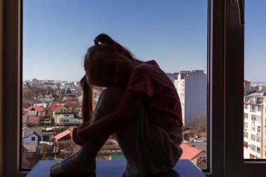 Küçük bir kızın silueti. Kafası dizlerinin üzerinde pencerenin arka planına yaslanmış.