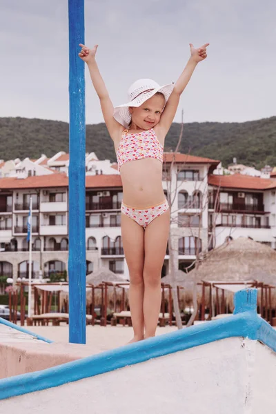 Little girl in swimsuit and beach hat stands on boat raising her hands up against the background of hotels and green hills