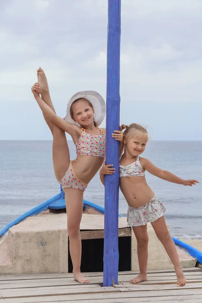 Mayolu iki küçük kız, beyaz ve mavi bir teknede, kumların üzerinde, arka planda fotoğraf için poz veriyorlar. Bir tanesi bacağını kaldırdı..