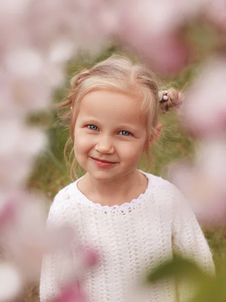 Blonde little girl in a white blouse in focus on a background of grass photographed through the flowers