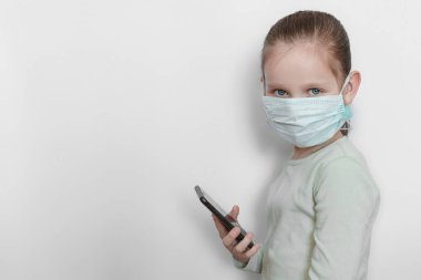 Elinde telefon ve yüzünde beyaz gri bir arka planda tıbbi maske olan açık renk monokrom süveter giyen küçük üzgün kız kameraya bakar.